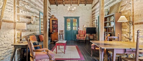 A rustic interior with exposed stone walls, wooden beams, a wooden table, a television, and a bookshelf.