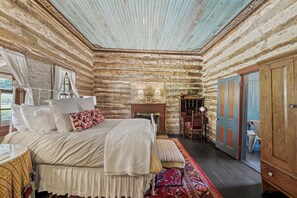 A log cabin bedroom with a bed, bedside table, lamp, and a view of the outside. - An 1865 cabin, home to four generations (Fredericksburg)