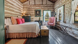 Ein Schlafzimmer in einer Blockhütte mit einem Bett, einem Sessel, einer Kommode und einem Fenster mit Vorhängen.