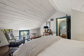 Een slaapkamer met een schuine kap, een bed, een stoel, een tafel en een deur naar een andere kamer.