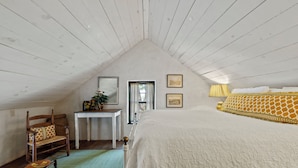 Ein Schlafzimmer mit schrägem Dach, einem Bett mit gemustertem Überwurf, einem Stuhl mit Sitzgeflecht, einem kleinen Tisch mit Dekoration und zwei gerahmten Bildern an der Wand.