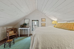 A bedroom with a sloped ceiling, a bed with a patterned bedspread, a chair with a woven seat, a small table with decorative items, and two framed pictures on the wall. - An 1865 limestone barn with gun-slot windows (Fredericksburg)