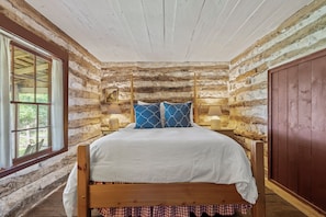 A rustic log cabin bedroom with a wooden bed, two bedside lamps, and a large window. - An 1847 cabin, among the area's oldest homes (Fredericksburg)