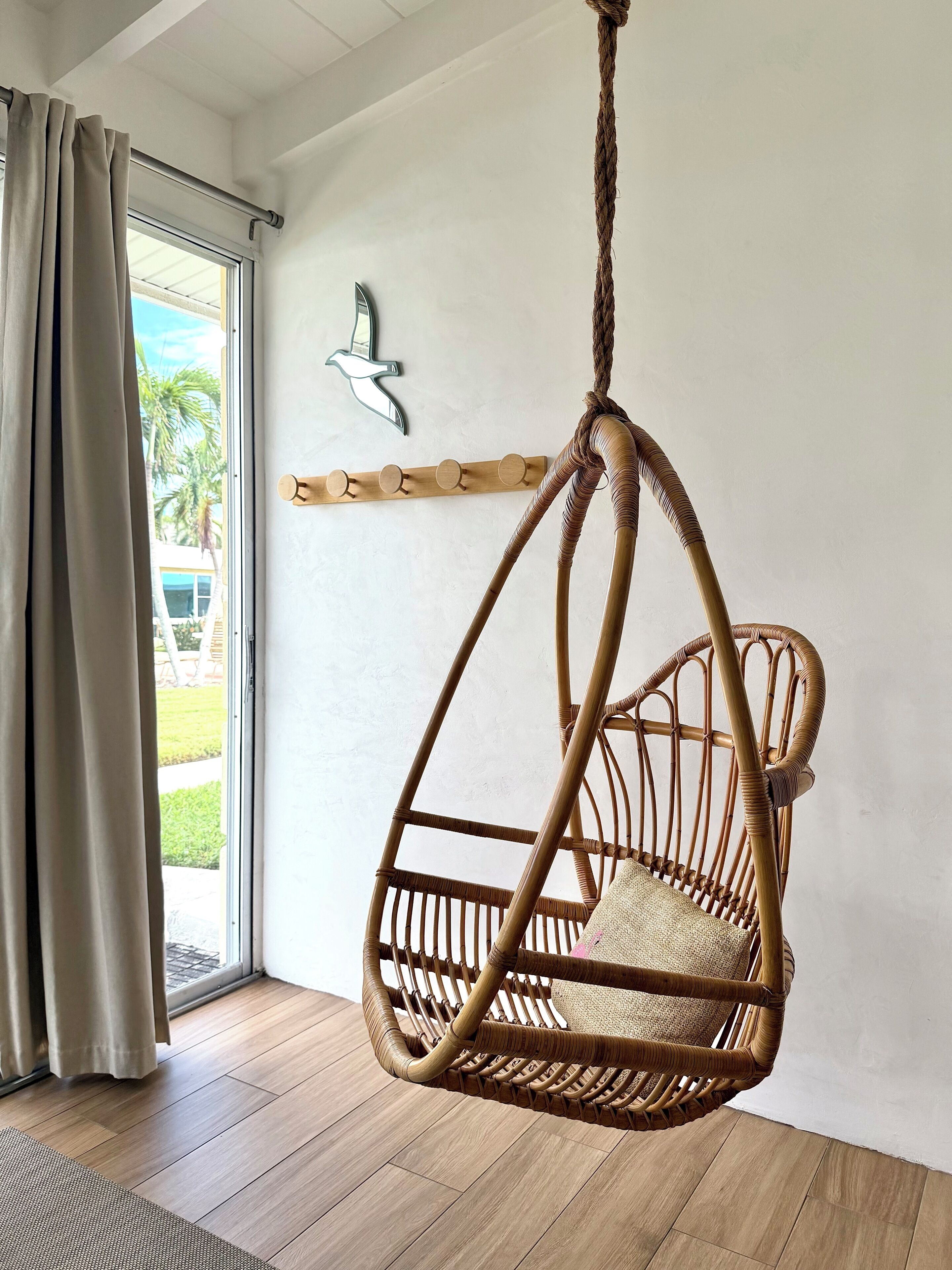 Un fauteuil suspendu en osier avec un coussin, un porte-manteau en bois et une fenêtre avec des rideaux.