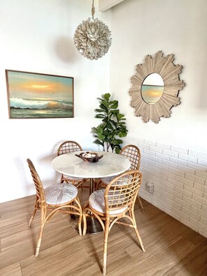 A dining area with a round table, wicker chairs, a potted plant, a wall-mounted mirror, and a framed painting.