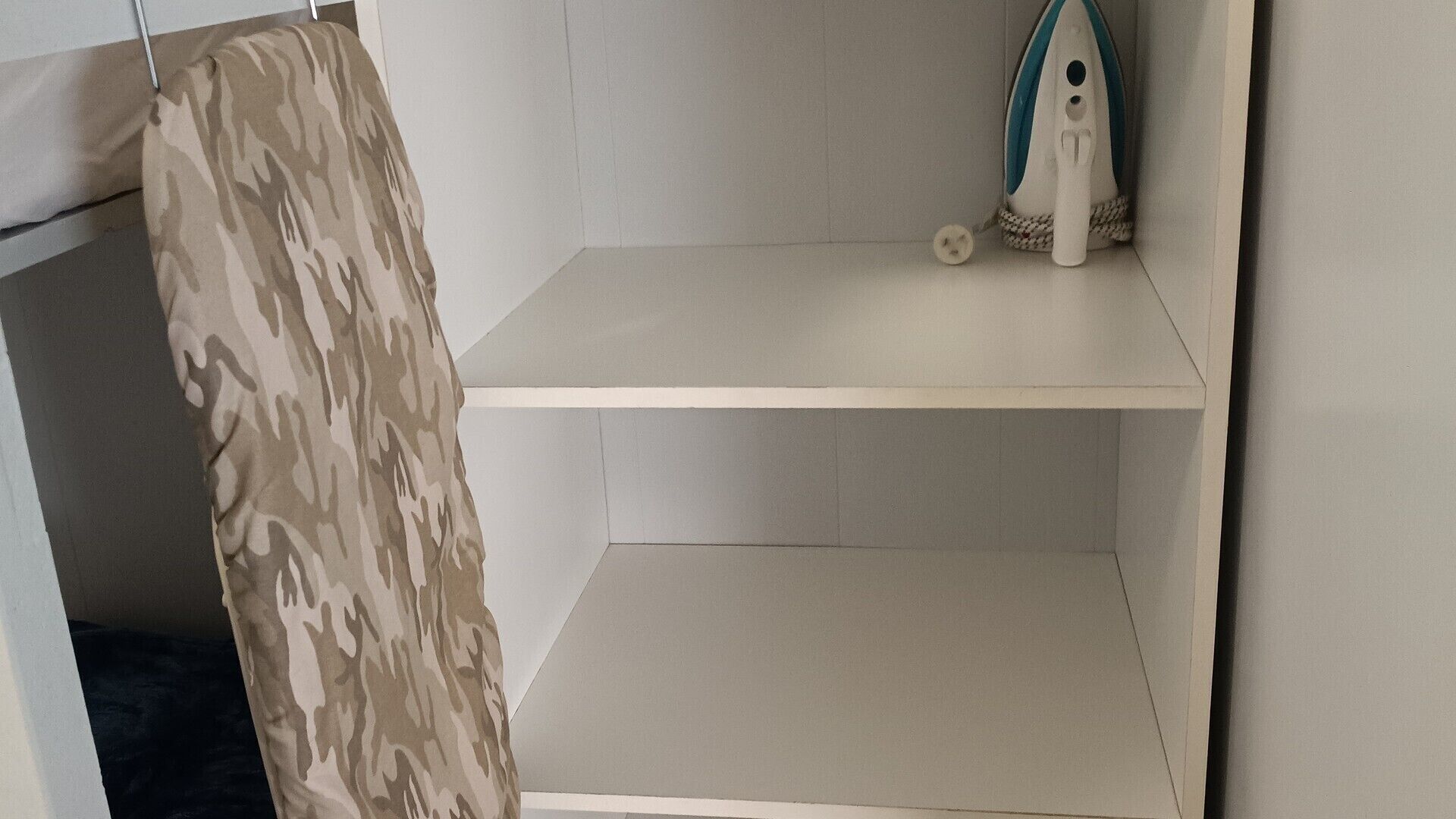 A corner of a room with a camouflage-patterned item hanging on the wall, a white shelf with a small object on it, and a white wall.