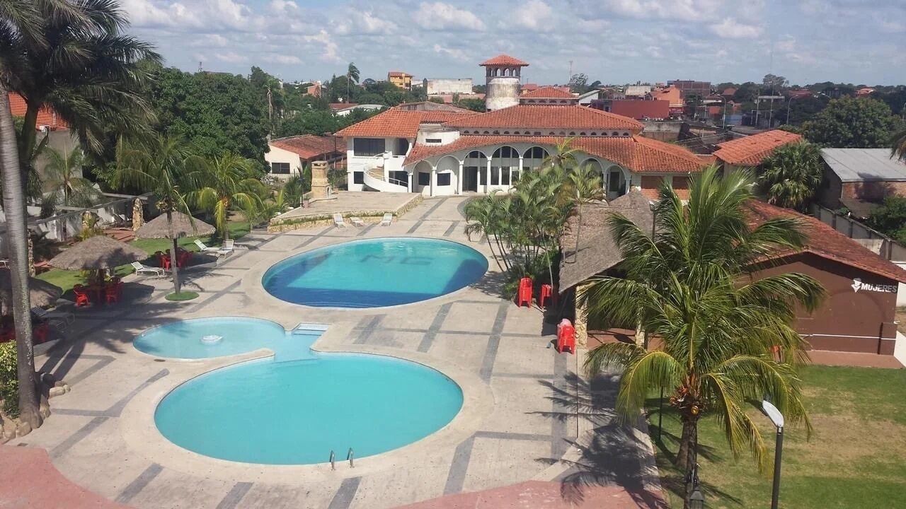 Un resort con diverse piscine, palme e vista su una cittadina con tetti rossi.