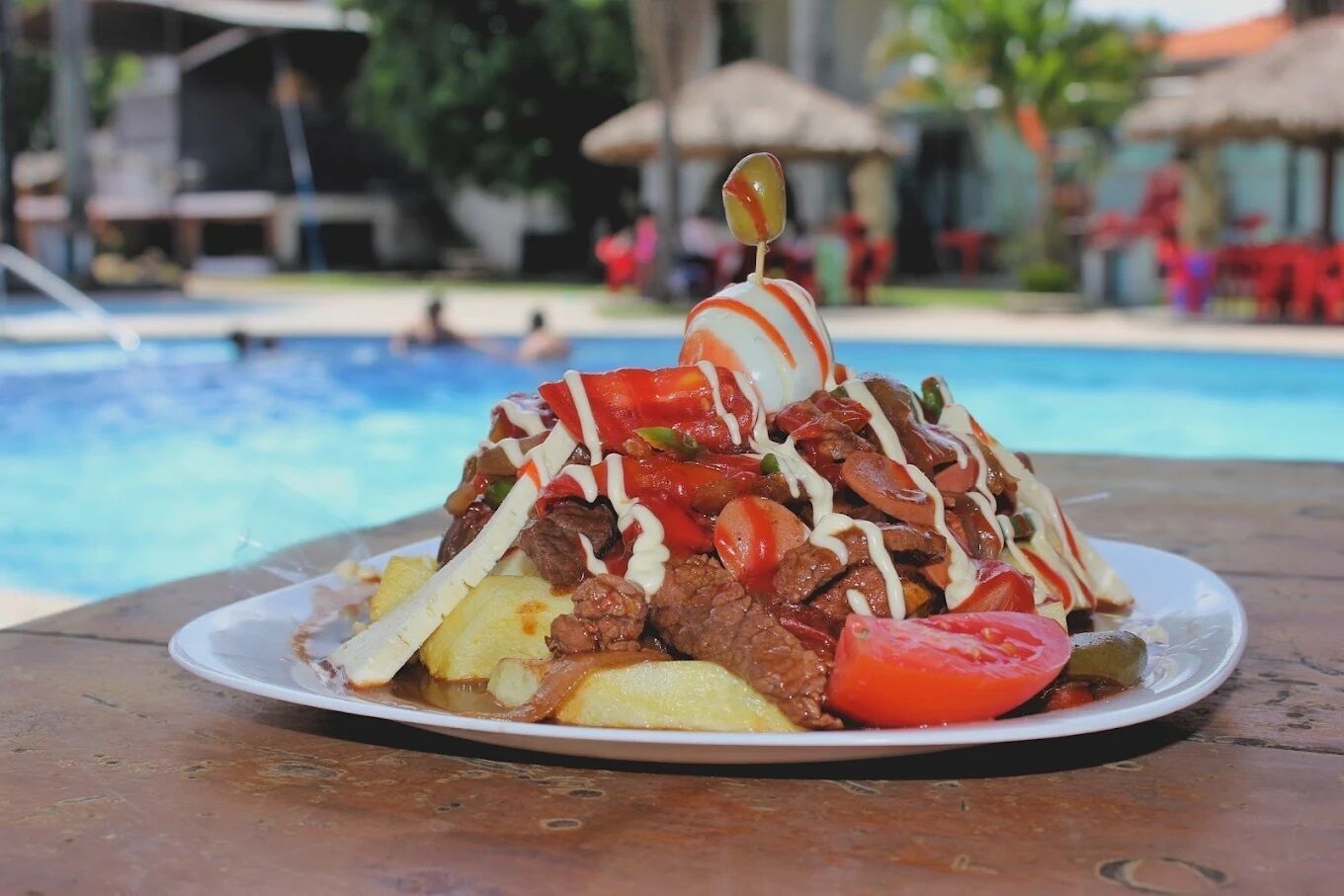 Un plato de comida con un guiso de carne, verduras y una rodaja de tomate sobre una mesa junto a una piscina.