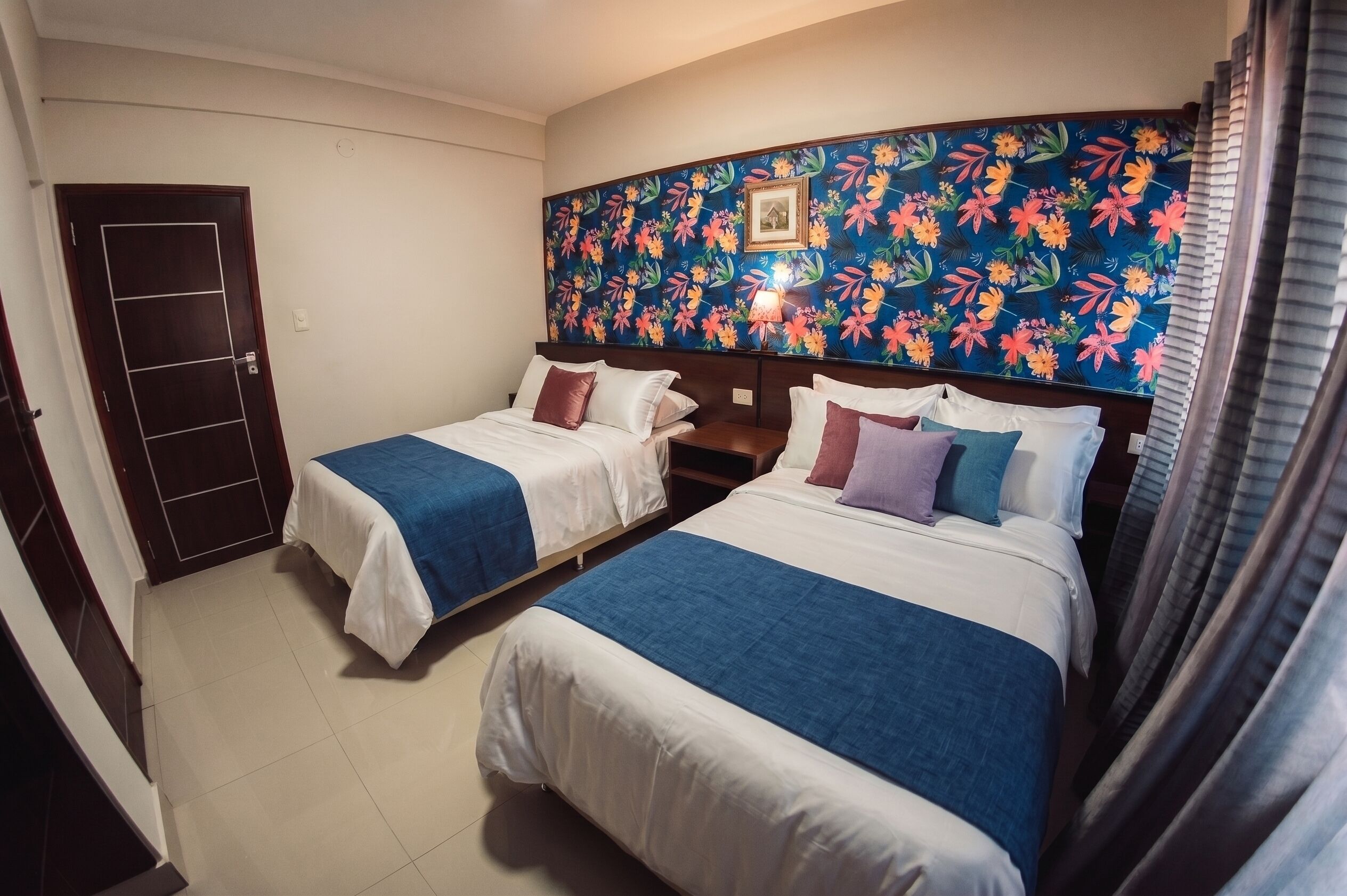Habitación de hotel con dos camas, cabecera estampada con motivos florales, una puerta y una ventana con cortinas.