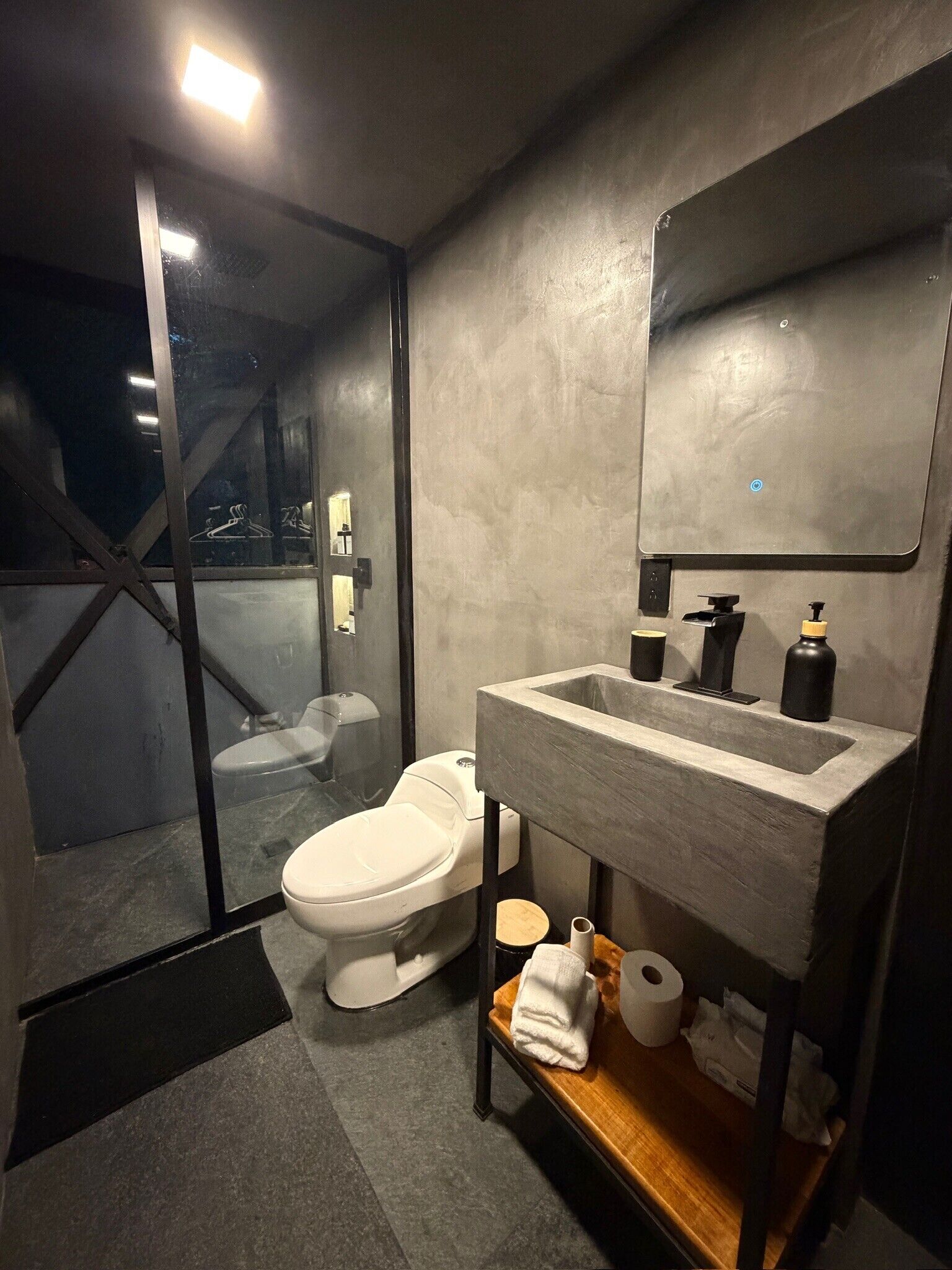 Et moderne badeværelse med brusekabine i glas, et hvidt toilet, en vask med et spejl over, og en hylde med toiletartikler.
