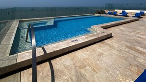 Uma piscina no terraço com água azul cristalina, rodeada por um deck de pedra e espreguiçadeiras azuis.
