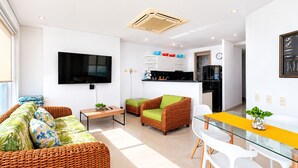 Uma sala de estar moderna com televisão de tela plana, móveis de vime, uma mesa de jantar de vidro e uma cozinha ao fundo.
