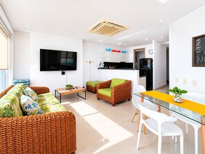 Ein modernes Wohnzimmer mit Flachbildfernseher, Rattanmöbeln, einem Glastisch und einer Küche im Hintergrund.