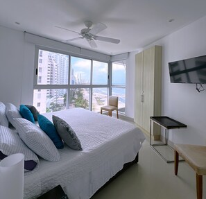 A modern bedroom with a large bed, a ceiling fan, a TV, and a view of the city through large windows. - 2-Bedroom Apartment in Bocagrande (Near the Historic Center) (Cartagena de Indias)