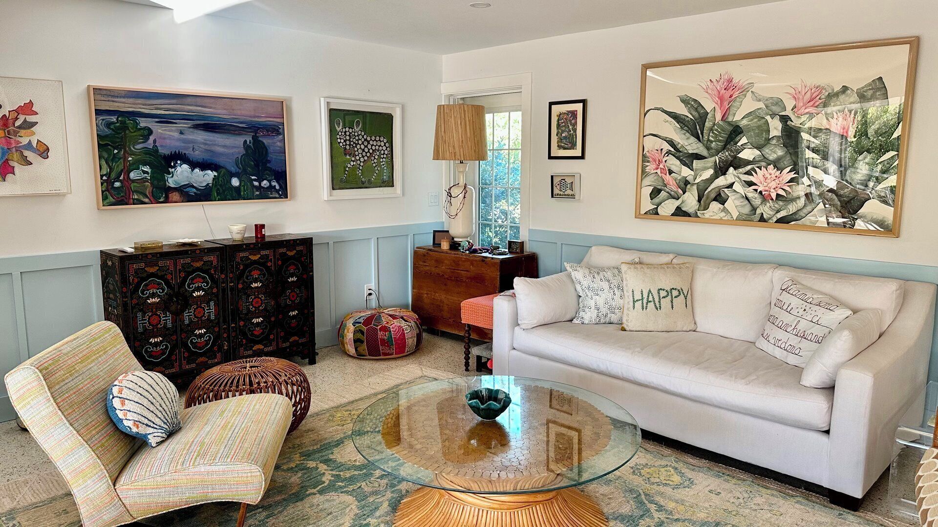 A living room with a white sofa, patterned armchairs, a glass coffee table, and various wall art.