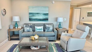 A living room with a sofa, a coffee table, a chair, and a painting of a seascape. - Sanibel Surfside 117 - Gulf front corner unit - All new - Sunrises & Sunsets & Gulf Views Galore! (Sanibel)
