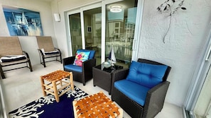 A sunlit patio with wicker furniture, a glass door leading to a balcony, and a painting on the wall. - Sanibel Surfside 117 - Gulf front corner unit - All new - Sunrises & Sunsets & Gulf Views Galore! (Sanibel)