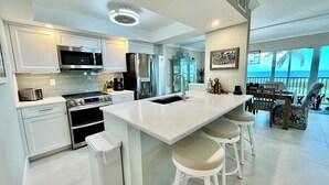 A modern kitchen with white cabinetry, stainless steel appliances, and a large island with bar stools. - Sanibel Surfside 117 - Gulf front corner unit - All new - Sunrises & Sunsets & Gulf Views Galore! (Sanibel)