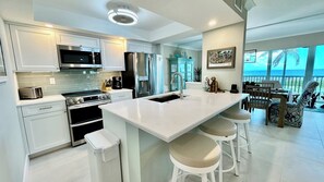 A modern kitchen with white cabinetry, stainless steel appliances, and a large island with bar stools. - Sanibel Surfside 117 - Gulf front corner unit - All new - Sunrises & Sunsets & Gulf Views Galore! (Sanibel)