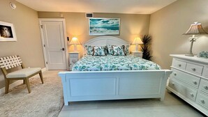 A bedroom with a bed, a chair, a dresser, a door, and a wall-mounted picture. - Sanibel Surfside 117 - Gulf front corner unit - All new - Sunrises & Sunsets & Gulf Views Galore! (Sanibel)