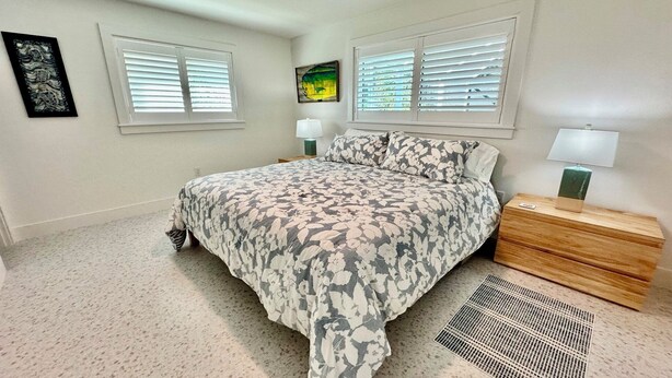 Um quarto com cama, duas mesas de cabeceira, um criado-mudo, janela com persianas e uma obra de arte pendurada na parede.