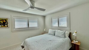 Ein Schlafzimmer mit einem Bett, zwei Nachttischlampen, einem Deckenventilator und zwei Fenstern mit Jalousien.