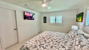 Ein Schlafzimmer mit Bett, Fernseher, einem Fenster mit Jalousien und einem Deckenventilator.