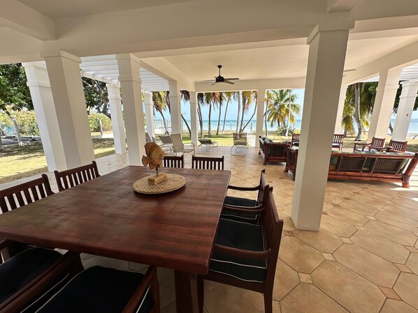 A covered outdoor dining area with a wooden table and chairs, a ceiling fan, and a view of the beach and ocean.