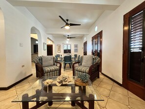 Uma sala ampla com uma mesa de centro de vidro, dois sofás estampados, uma mesa de jantar com cadeiras e um ventilador de teto.