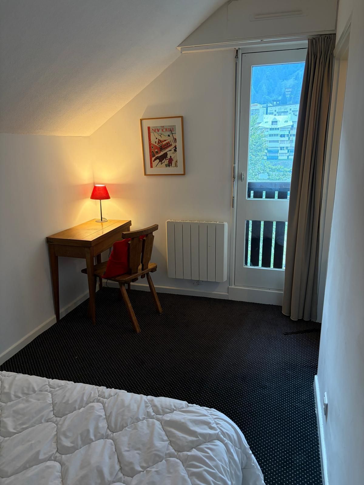 A hotel room with a bed, a desk with a lamp, a radiator, and a window with a view of buildings.