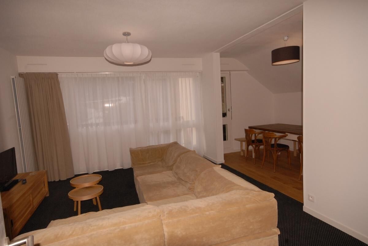 A living room with a beige sofa, two small round tables, a TV, and a dining area with a table and chairs.