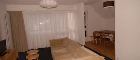 A living room with a beige sofa, two small round tables, a TV, and a dining area with a table and chairs.