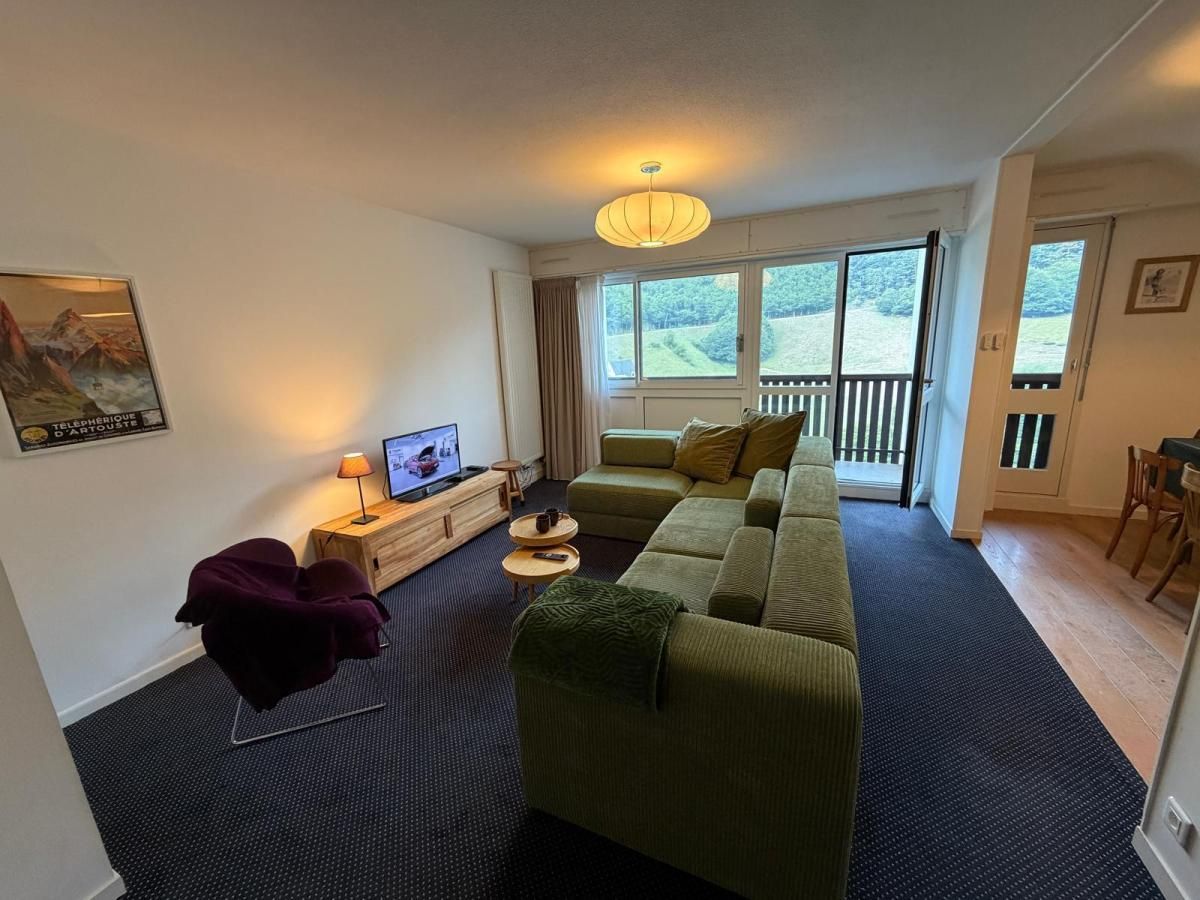 A living room with a sofa, a television, a small table with a lamp, and a window with a view of greenery.