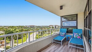 Ein Balkon mit zwei Stühlen und einem Tisch, der auf eine grüne Landschaft und Gebäude blickt.