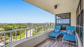 A balcony with two chairs and a table, overlooking a green landscape and buildings. - Remodeled Beachfront Corner Condo | Marco Island | Sleeps 6 (Marco Island)
