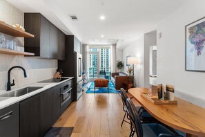 A modern kitchen with dark cabinets, a stainless steel sink, and a wooden dining table with a blue rug. - Luxury Downtown Stay l Rooftop Pool & Skyline Views (Austin)