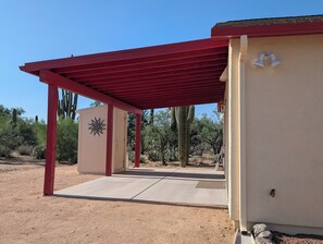 Un espace extérieur abrité, doté d’un toit rouge, d’une porte ornée d’un motif solaire et offrant une vue sur des cactus et des arbres.