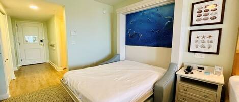 A hotel room with a bed, a desk with a chair, a nightstand, a wall-mounted TV, and a door leading to another room.