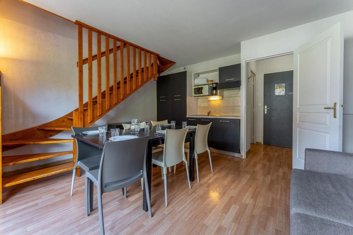A modern kitchen with dark cabinets, a wooden staircase, and a dining area with a table and chairs.