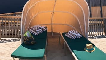 Une tente installée sur la plage, avec deux chaises longues vertes, chacune agrémentée d’une serviette et d’un chapeau.