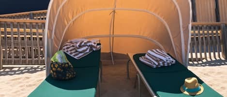A beachside tent with two green lounge chairs, each with a towel and a hat.