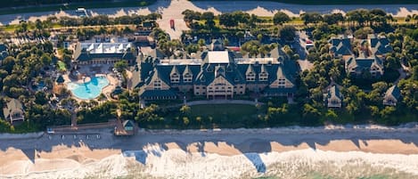 An aerial view of a resort with a large building, a swimming pool, and a beach.