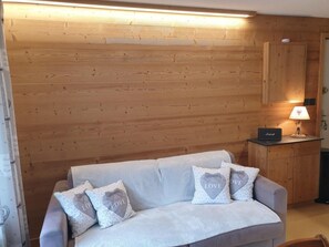 A room with a wooden wall, a grey sofa with heart-shaped pillows, a small cabinet, and a lamp. - 120m from ski slopes, 2nd floor, balcony, parking, tv, ski locker, 28m², Valloire (Valloire)