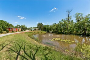 A serene lakeside setting with a clear sky, a small pond with lily pads, and a pathway leading to a row of cabins. - Modern Boxabl Casita | Stillwater OK | Pasadera 7 (Stillwater)