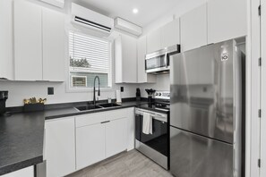 A modern kitchen with white cabinetry, a stainless steel refrigerator, a built-in oven, and a window with blinds. - Modern Boxabl Casita | Stillwater OK | Pasadera 7 (Stillwater)
