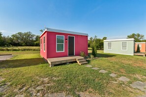 A small pink mobile home with a wooden deck and a green door, situated in a grassy area with trees in the background. - Modern Boxabl Casita | Stillwater OK | Pasadera 7 (Stillwater)