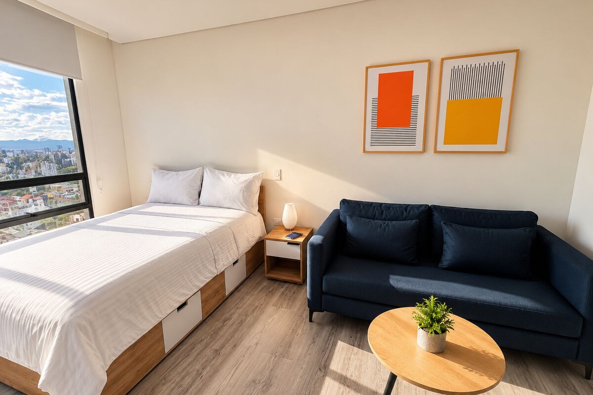 Quarto de hotel moderno com cama, sofá, mesinha com planta e duas obras de arte emolduradas na parede.
