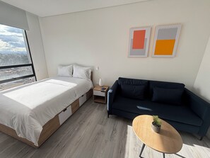 Un dormitorio moderno con cama, un sofá oscuro, una mesita con una planta y obras de arte en la pared.