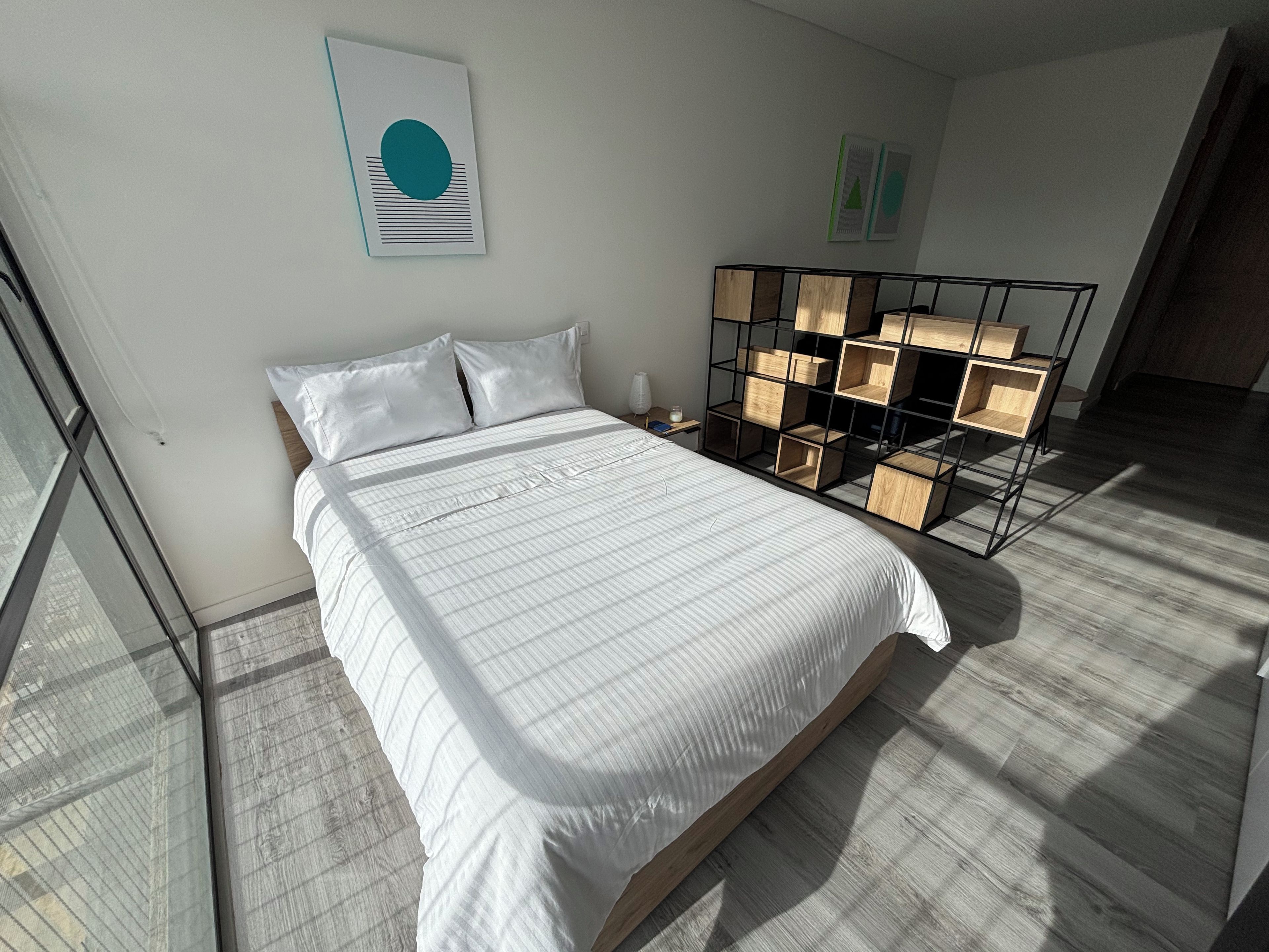 Un dormitorio moderno con una cama grande, una mesita de noche de madera, una estantería en la pared y un ventanal amplio.