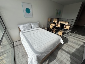 Quarto moderno com cama grande, mesa de cabeceira de madeira, estante fixada na parede e uma janela ampla.